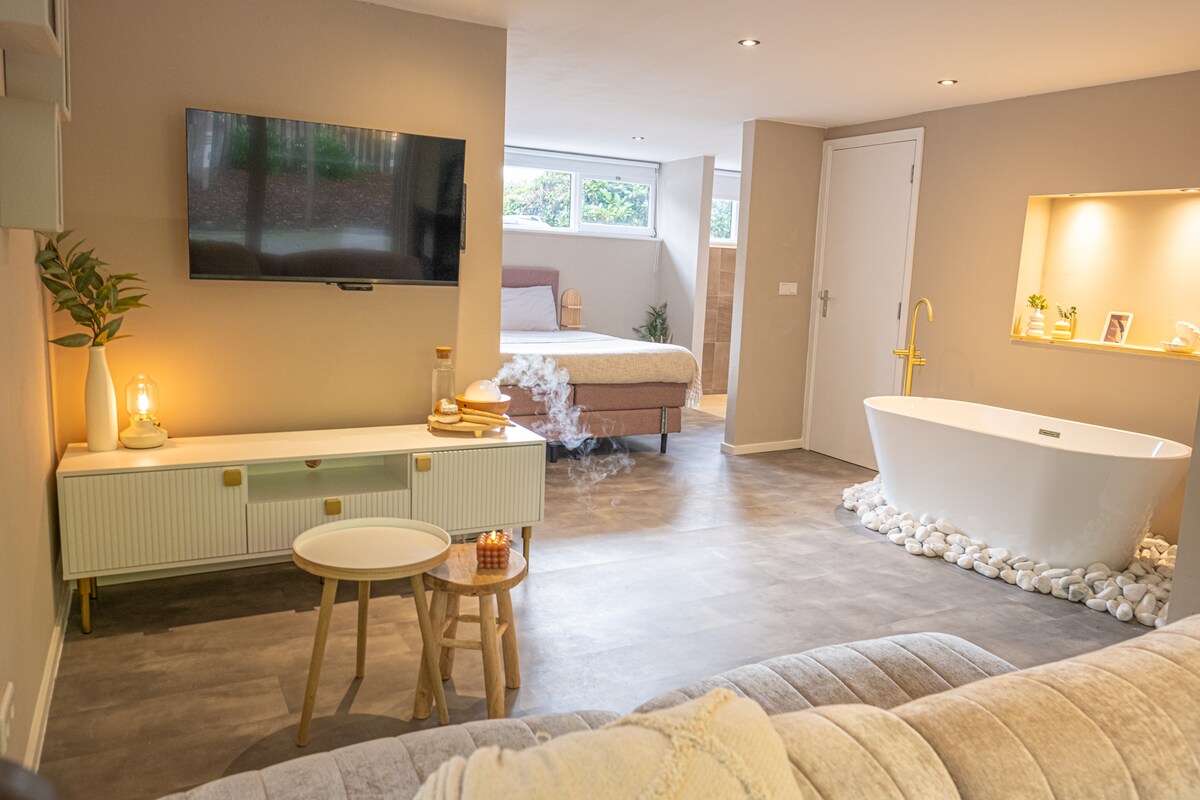 A spacious living area is featured with a comfortable sofa and a television mounted on the wall. Natural light enters through windows, illuminating the freestanding bathtub positioned on pebbled flooring. A muted color palette is present throughout, enhancing the serene ambiance.