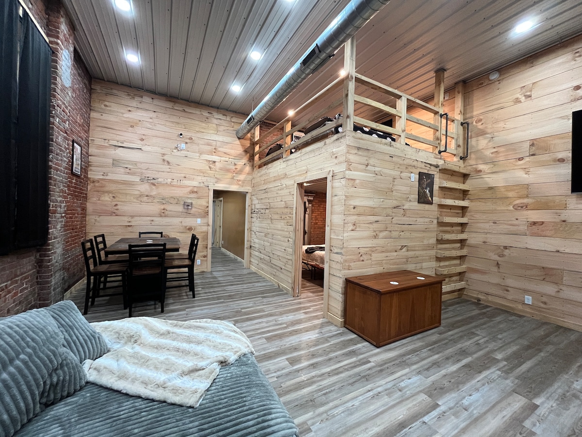 An open living area is highlighted by wooden walls and a loft above. A spacious sofa with a soft throw blanket is present in the foreground, while a dining table and chairs are positioned nearby. Natural light enhances the warm tones of the wood.