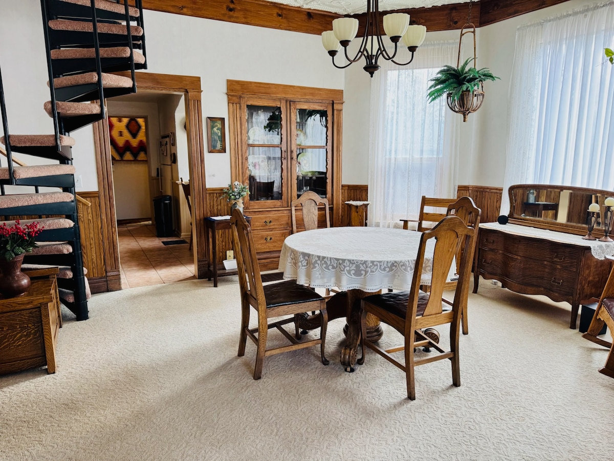 A spacious dining area features a round table surrounded by wooden chairs, enhanced by a lace tablecloth. A large cupboard is visible, complemented by warm wood tones throughout. Natural light enters through large windows with sheer curtains, while tasteful decor accents the room's charm.