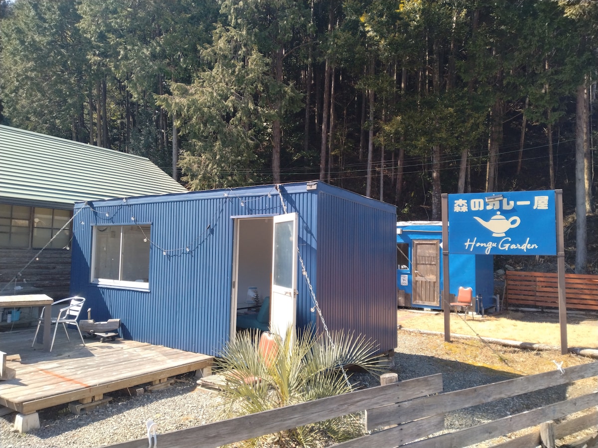 A compact blue container house is surrounded by lush woods, featuring a small porch with seating. A wooden deck is situated in front of the house, contributing to a natural outdoor setting. The entrance is framed by a welcoming sign indicating 'Hongo Garden.'