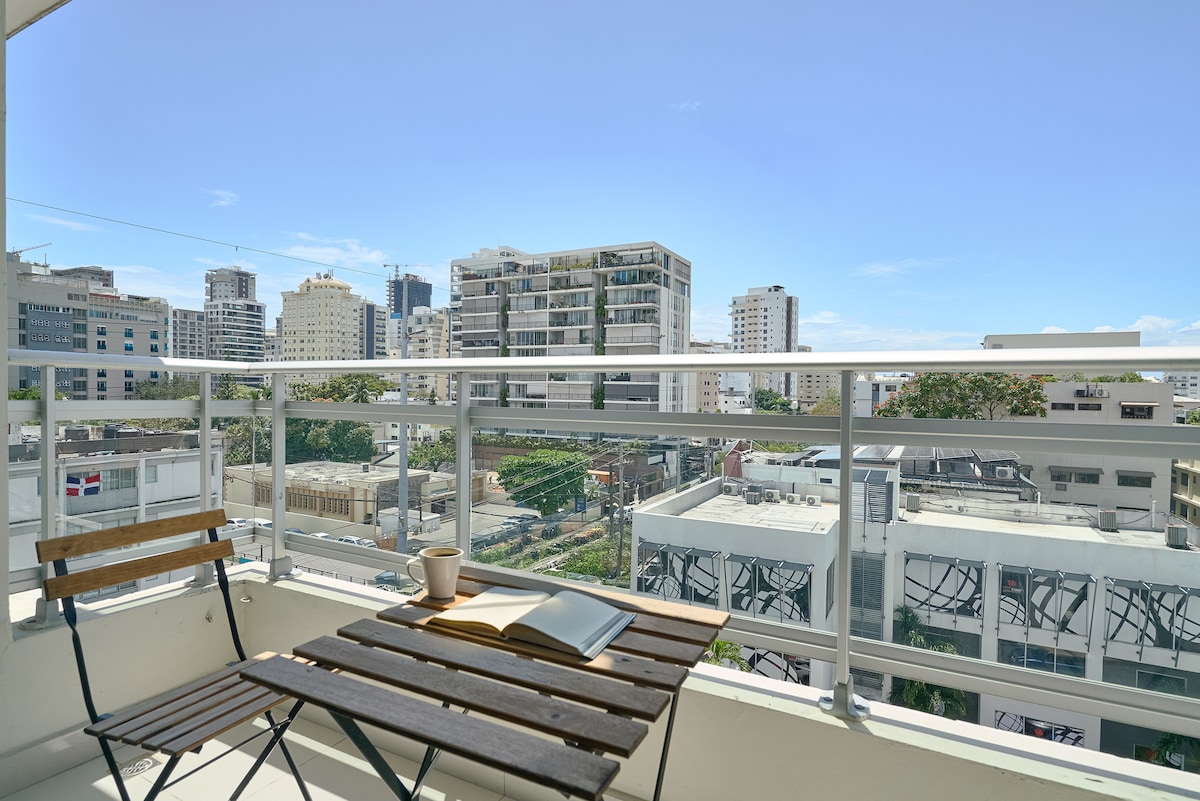 A balcony view captures a bustling urban landscape, with modern buildings visible in the distance. A small wooden table and two chairs are positioned on the balcony, inviting relaxation while enjoying the scenery. The sky is clear, enhancing the overall ambiance.