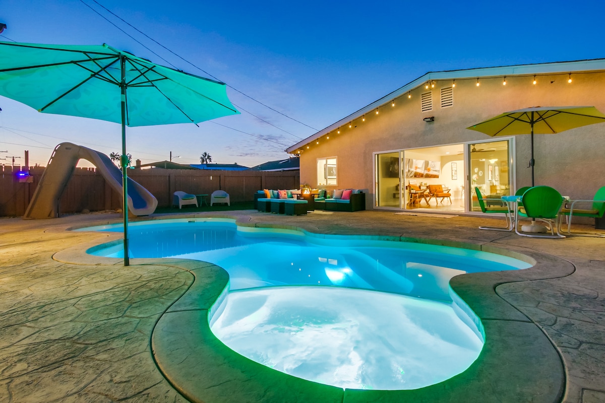 The outdoor area features a sparkling pool with a unique shape, illuminated in soft blue light. Brightly colored seating surrounds the pool, complemented by oversized umbrellas providing shade. In the background, a modern home is visible, with large glass doors that invite natural light into the living space.