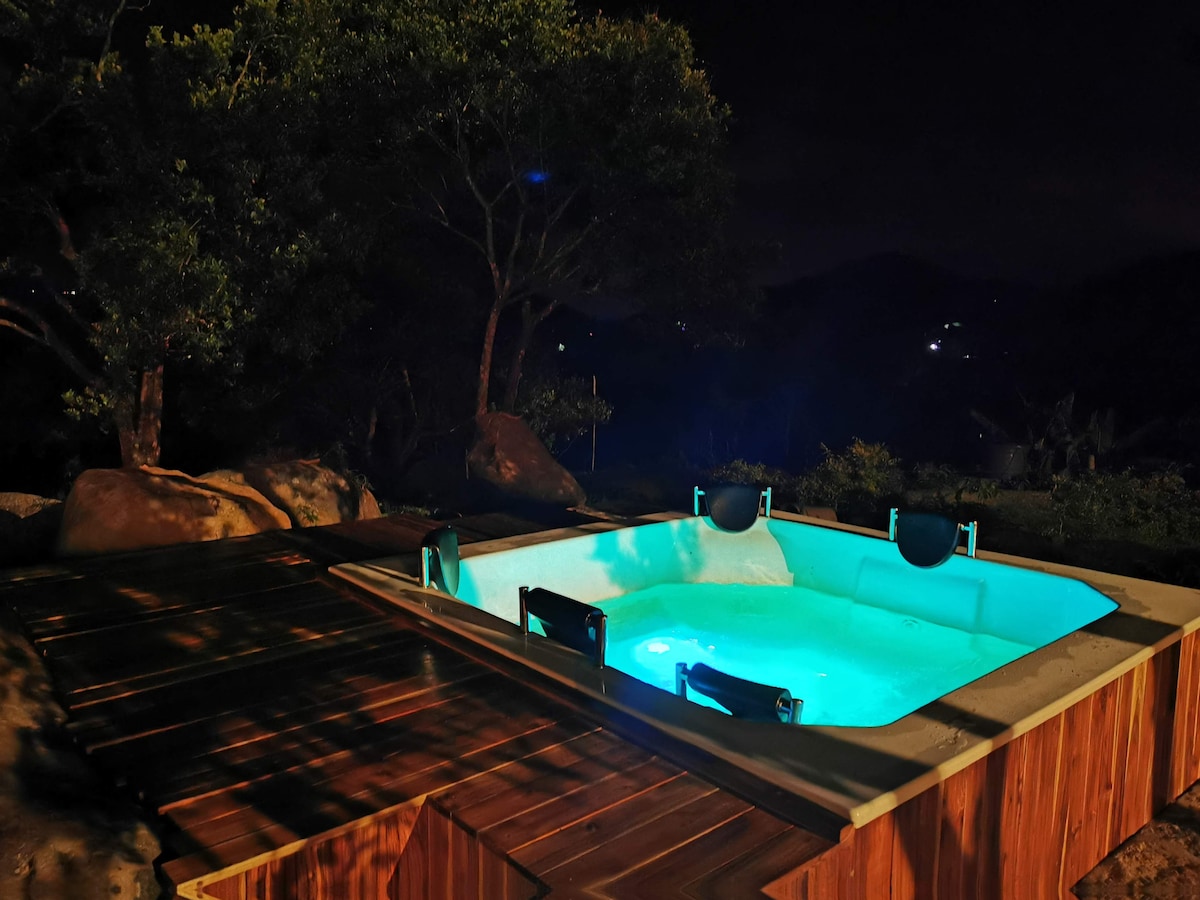 A private jacuzzi is set against a serene nighttime landscape, featuring soft blue lighting that creates a calming ambiance. The surrounding area includes wooden decking and vegetation, enhancing the tranquil feel of the outdoor space.