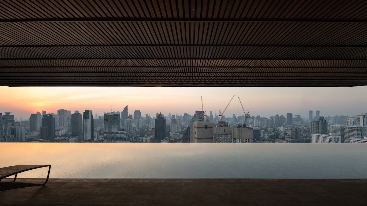 An infinity pool stretches across the foreground, offering a sweeping view of the Bangkok skyline at dusk. The horizon features numerous high-rises and construction cranes, with warm hues reflecting off the water's surface. A lone lounge chair is positioned at the edge.