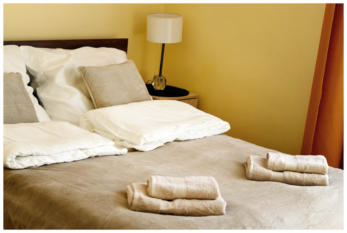 A well-appointed bedroom features a spacious bed with neatly arranged pillows and linens. Two folded towels are placed on the warm-toned bedspread. A bedside lamp provides soft lighting, complementing the gentle yellow wall color and inviting atmosphere.