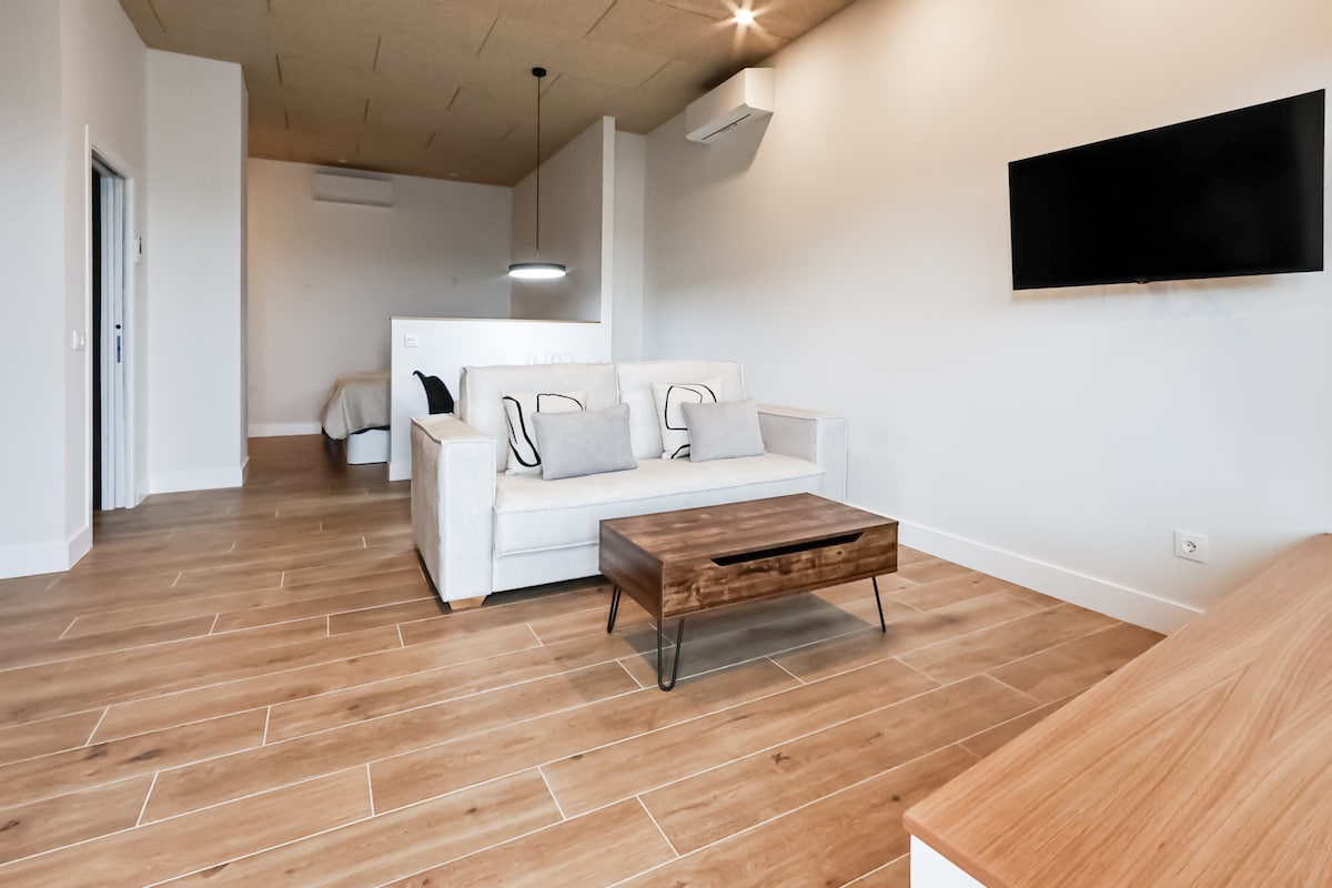 A spacious living area is presented with wood-style flooring and light-colored walls. A comfortable sofa faces a wall-mounted television, complemented by a simple wooden coffee table in front. Natural light filters in, creating a bright and inviting atmosphere.