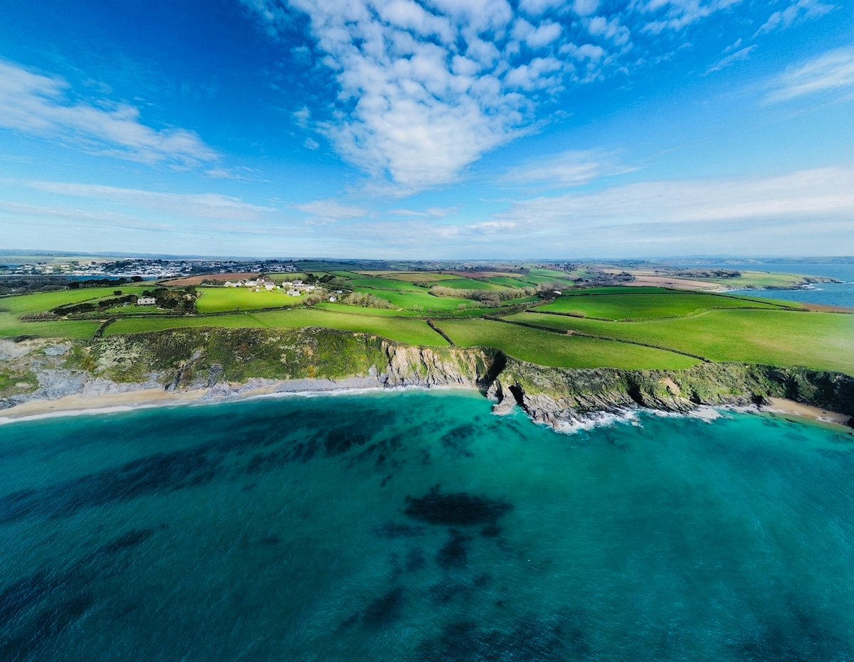 A wide coastal view captures the vibrant hues of the sea blending with the green landscape. The clear blue sky is dotted with soft clouds, while the shoreline features rocky cliffs meeting the tranquil waters, extending toward the horizon.