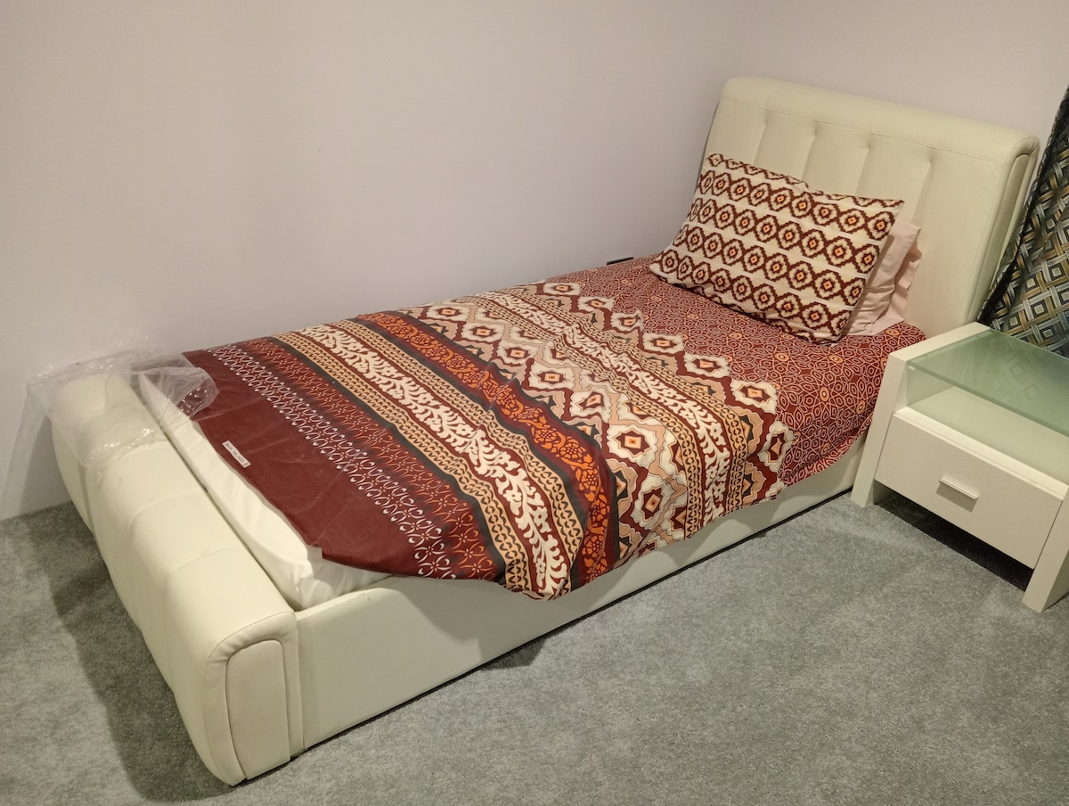 A spacious bed is positioned against a neutral wall, covered with a patterned blanket featuring earthy tones. A decorative pillow adds comfort. Beside the bed, a small nightstand can be seen, contributing to the room's functionality.