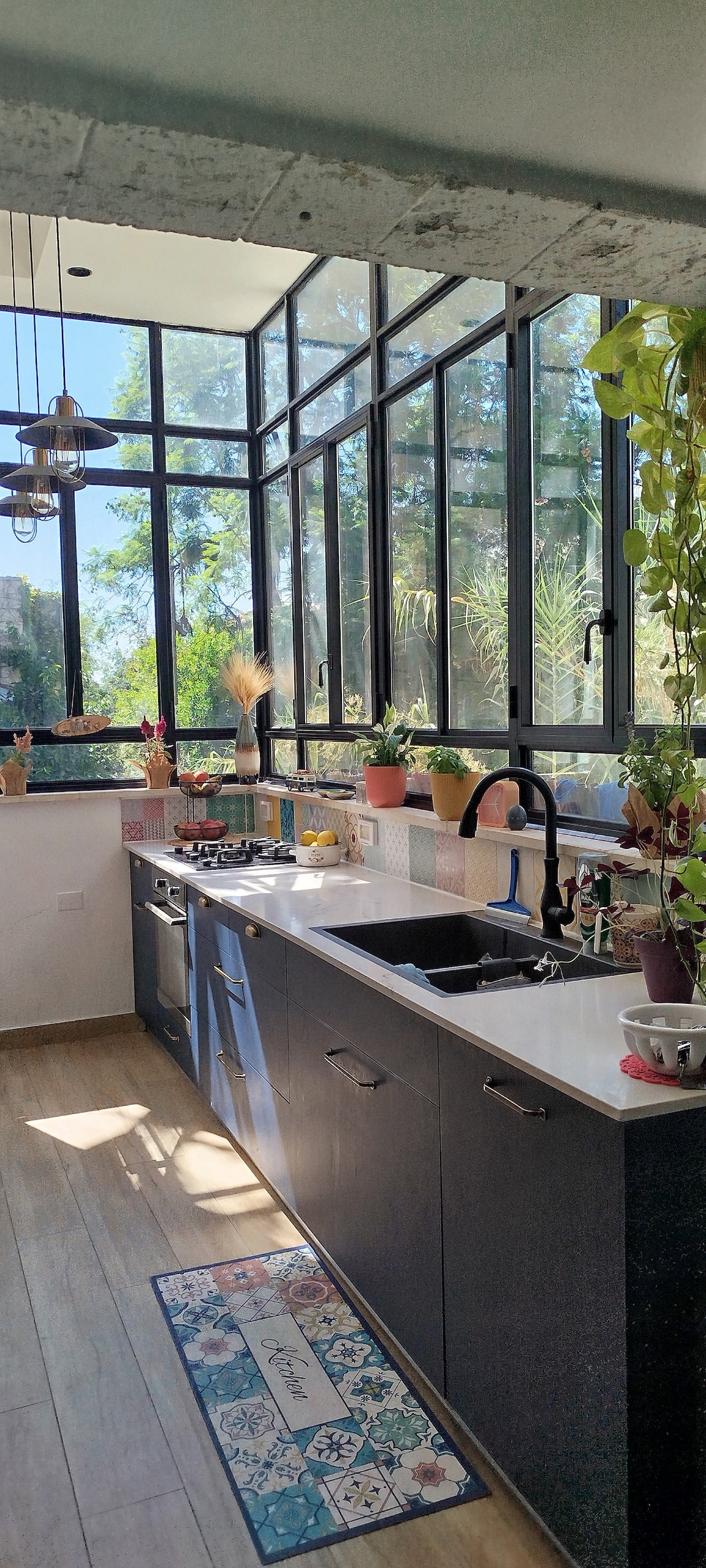 A well-lit kitchen is present, featuring large windows that provide ample natural light and a view of greenery. The space includes dark cabinetry, a white countertop, and a variety of potted plants. A sink and stovetop are positioned along the countertop, enhancing functionality.
