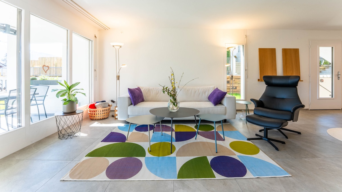 The living room features a modern sofa with decorative cushions, complemented by a circular coffee table. A colorful area rug with large geometric patterns adds interest to the space. Natural light streams in through large windows, illuminating plants placed by the glass doors.