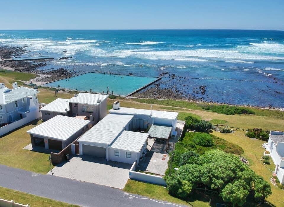An aerial view captures a spacious beach house positioned near the ocean. The property features well-maintained gardens, a swimming pool, and direct access to the tidal pool nearby. Waves gently lap against the shore in the background, with coastal vegetation framing the scene.