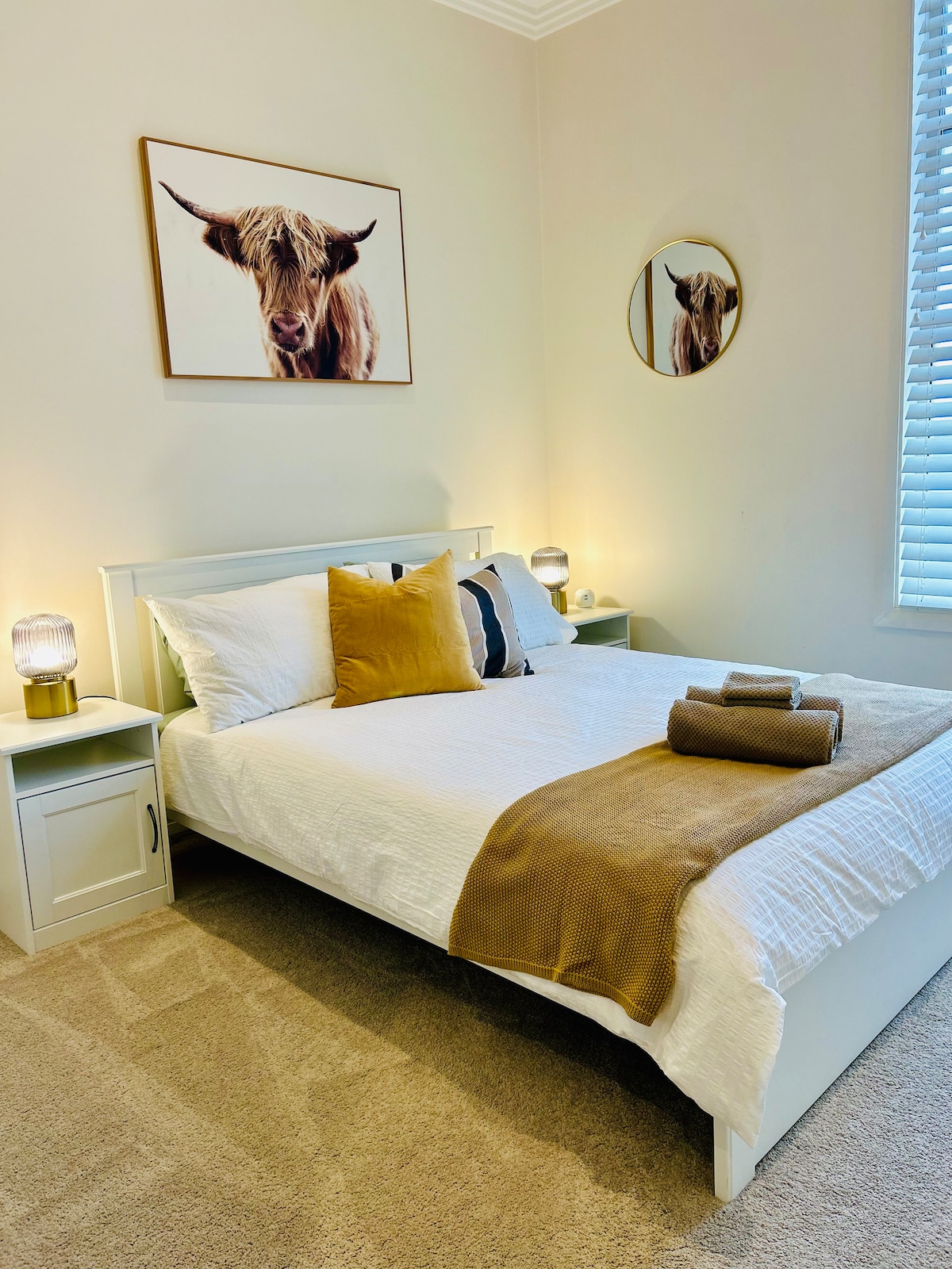 A bedroom is presented with a bright, inviting ambiance, featuring a spacious bed topped with neutral-toned linens and decorative pillows. Soft lighting from bedside lamps softly illuminates the room, complemented by a wall-mounted circular mirror and framed artwork above the bed.