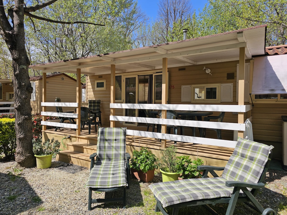 An outdoor area features a spacious wooden deck with white railings. Two lounge chairs are positioned in relaxed seating. A dining table and chairs are visible, surrounded by greenery, with a tranquil ambiance created by the surrounding trees.