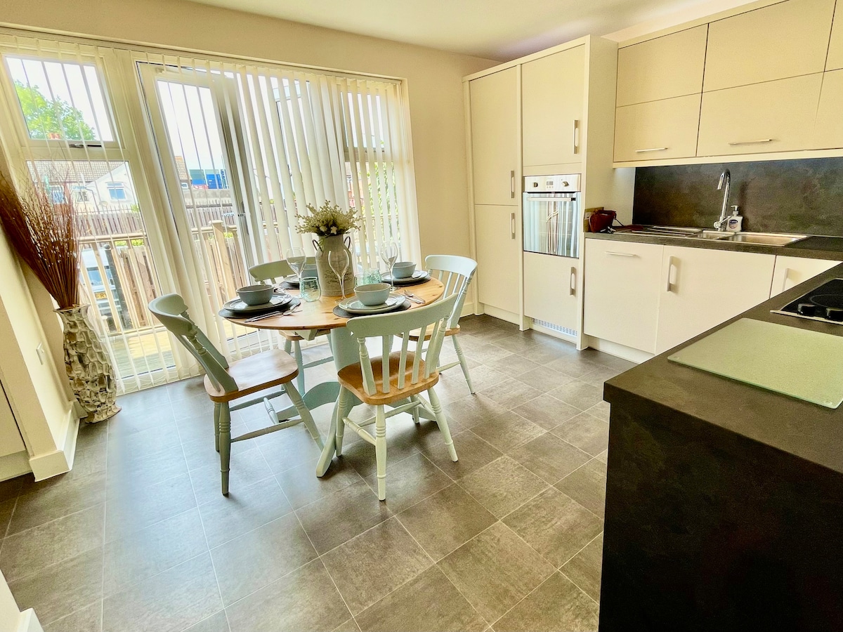 A modern kitchen area is featured, showcasing a round wooden dining table set for four. Light-colored chairs surround the table, and large sliding doors allow natural light to fill the space. Sleek cabinetry and dark countertops provide a contemporary backdrop.