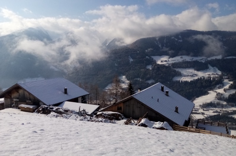 Snow-covered roofs of wooden houses are visible amidst a wintry landscape. Rolling hills and mountains create a serene backdrop, with patches of snow and evergreen trees. Soft clouds drift across a bright blue sky, enhancing the peaceful, natural environment.