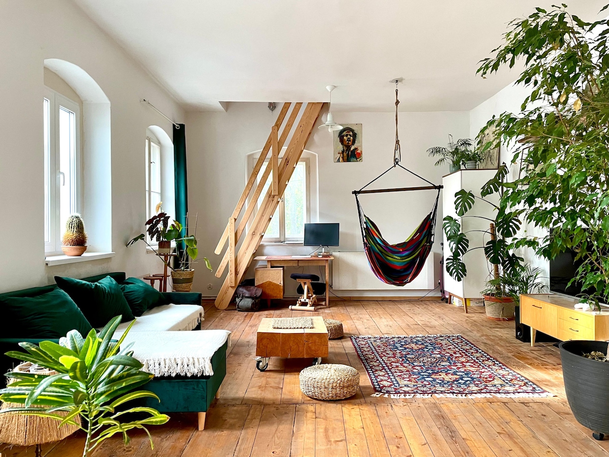 A bright living area features a combination of green sofas and a decorative rug over wooden flooring. A colorful hammock is suspended from the ceiling, accompanied by houseplants throughout the room. A desk with a computer is positioned near a window, providing natural light.