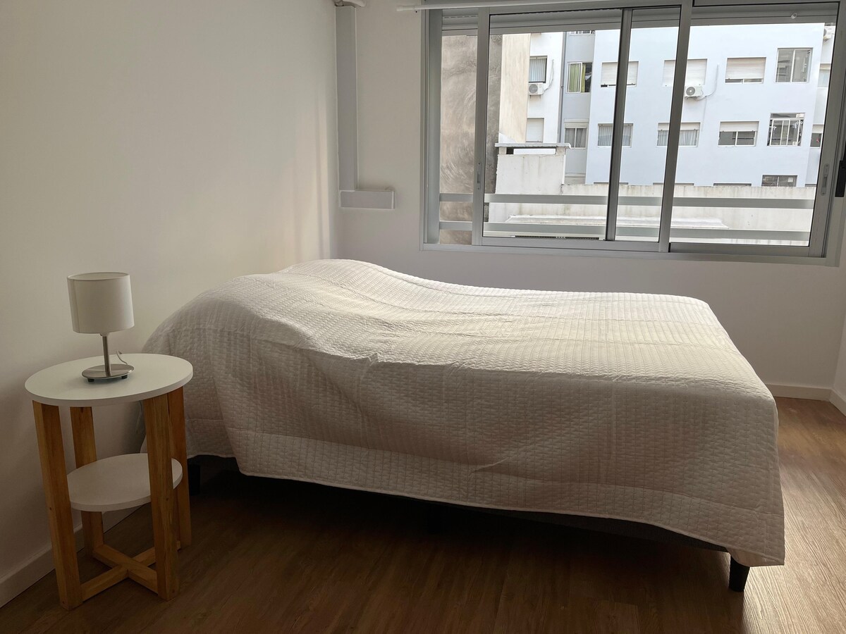 A spacious bed is covered with a light quilt, positioned near large windows that allow natural light to fill the room. A small bedside table with a lamp stands beside the bed, providing a practical touch to the minimalist decor.