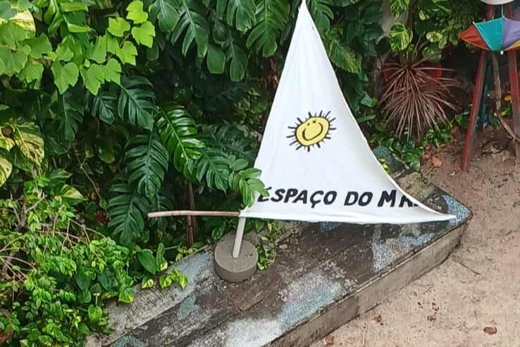 A small decorative sailboat flag is displayed in the garden, featuring a sun symbol and the words 'Espaço do Mar.' Lush greenery surrounds the flag, contributing to a vibrant natural setting. The flag stands on a sturdy base, complemented by sandy ground and additional tropical plants nearby.