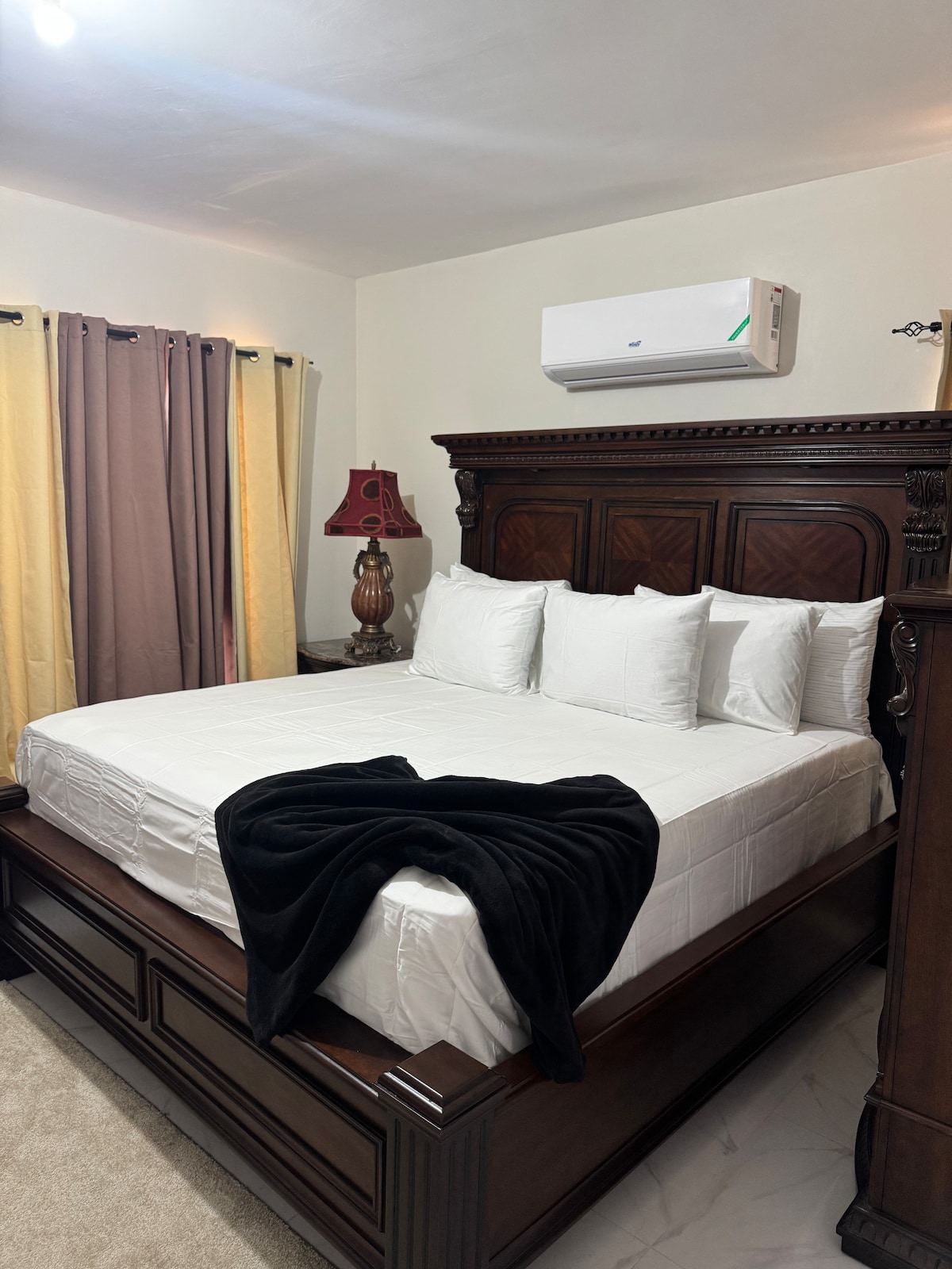 A well-furnished bedroom features a spacious bed adorned with white pillows and a dark throw blanket. A wooden nightstand holds a stylish lamp beside the bed. Large windows are dressed with light curtains, allowing for a blend of natural light and privacy.
