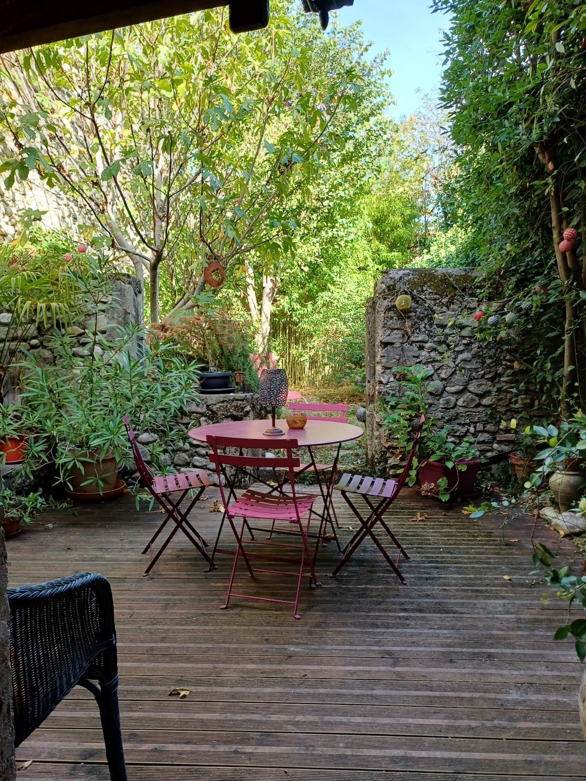 A charming outdoor seating area is shown, featuring a round table surrounded by four pink folding chairs. Lush greenery and potted plants provide a natural backdrop, alongside a stone wall. Sunlight filters through the trees, creating a serene atmosphere.
