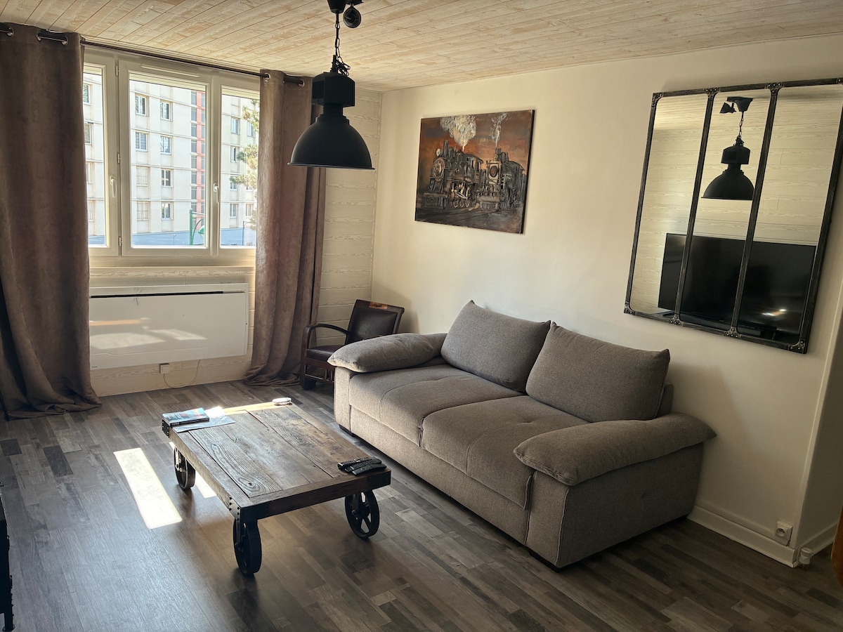 The living area features a comfortable gray sofa facing a mounted television. A rustic coffee table made of wood rests on the floor, complemented by a stylish light fixture overhead. Natural light enters through large windows covered by long curtains.