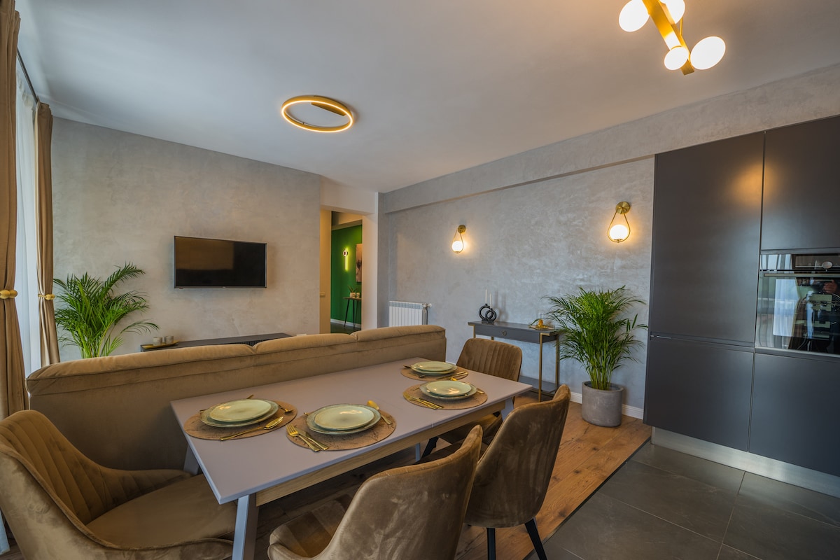 A modern dining area features a sleek table set for six, with neutral-toned plates and elegant glassware. Comfortable upholstered chairs surround the table. Soft lighting from overhead fixtures complements the space, alongside decorative plants that add a touch of greenery to the room.