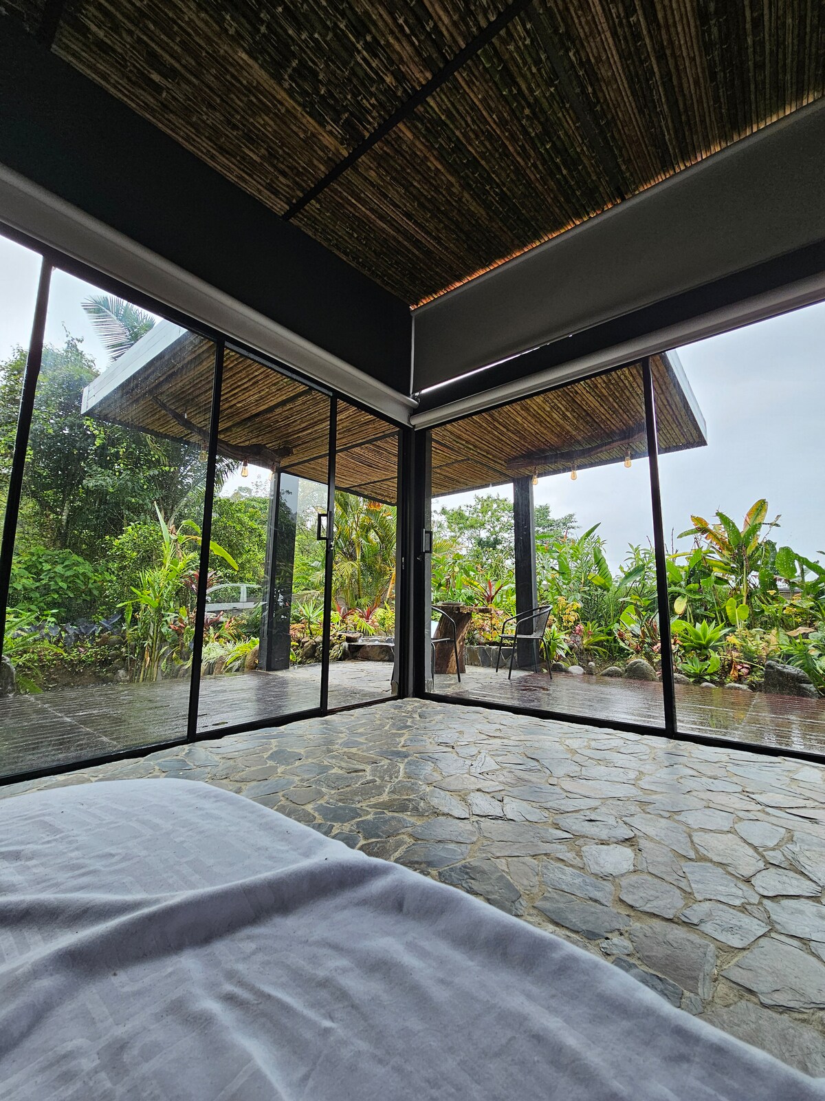 A spacious interior view reveals large glass windows that open to a lush tropical garden. Natural light floods the room, highlighting a stone floor and bamboo ceiling. Comfortable outdoor seating is visible on a wooden deck, surrounded by vibrant greenery and tropical plants.