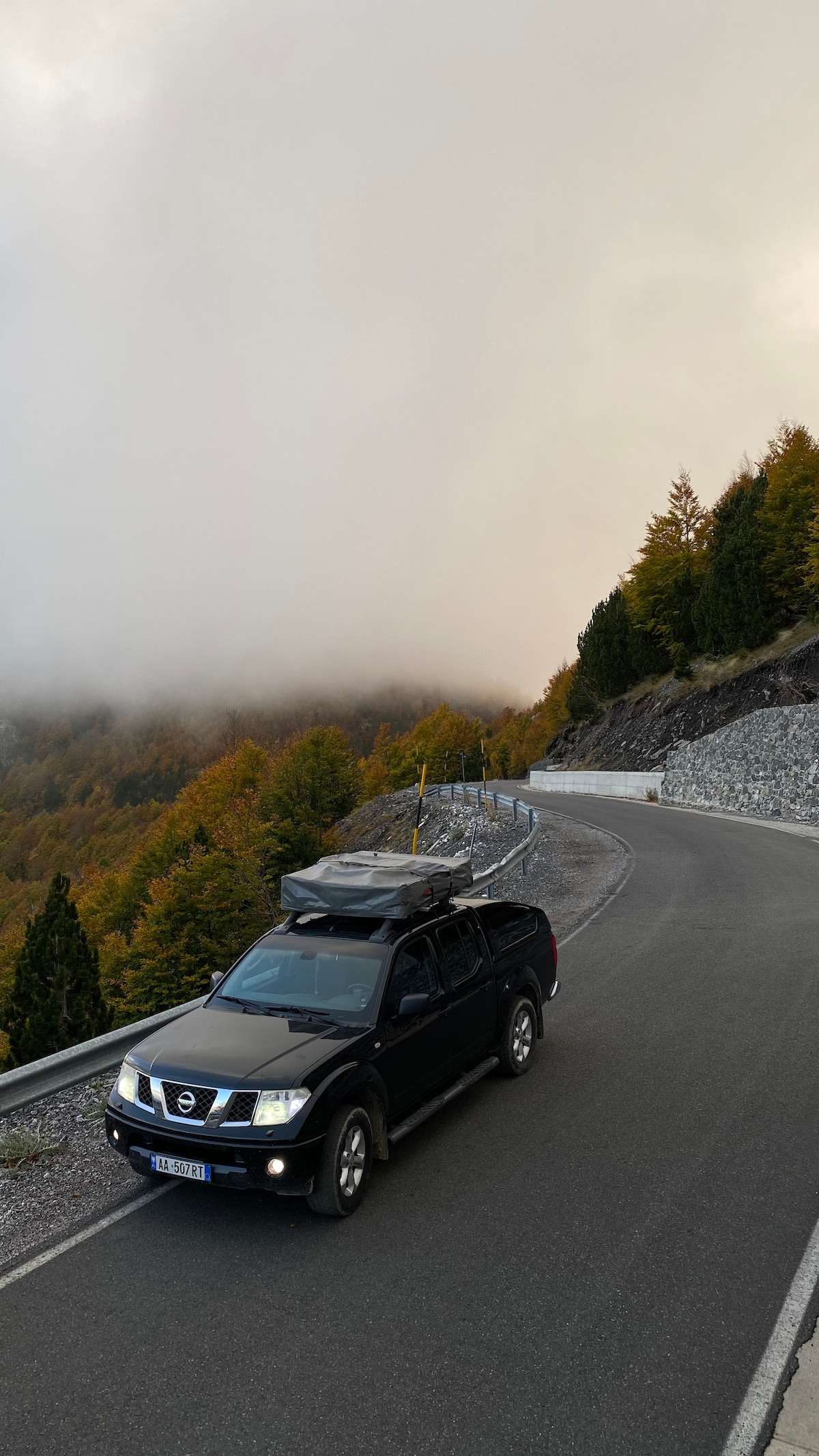 A black 4x4 vehicle is parked on a winding mountain road, surrounded by dense fog and autumn-colored trees. The road curves gently along the edge of a steep drop, offering a view of the natural landscape obscured by mist.
