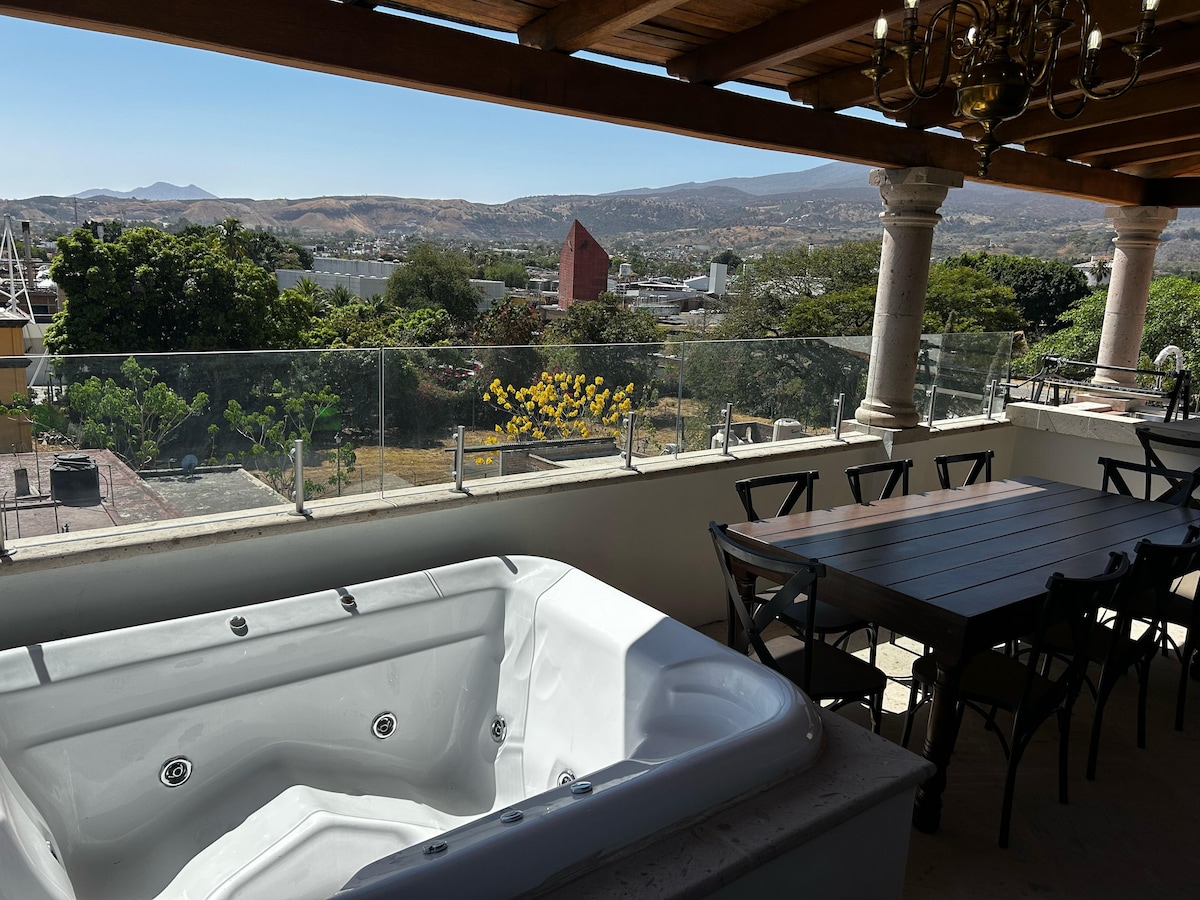 A spacious terrace features a white jacuzzi and a long dining table with dark chairs, set against a backdrop of rolling hills and vibrant greenery. Clear glass railings provide unobstructed views of the surroundings, allowing for a seamless outdoor experience.