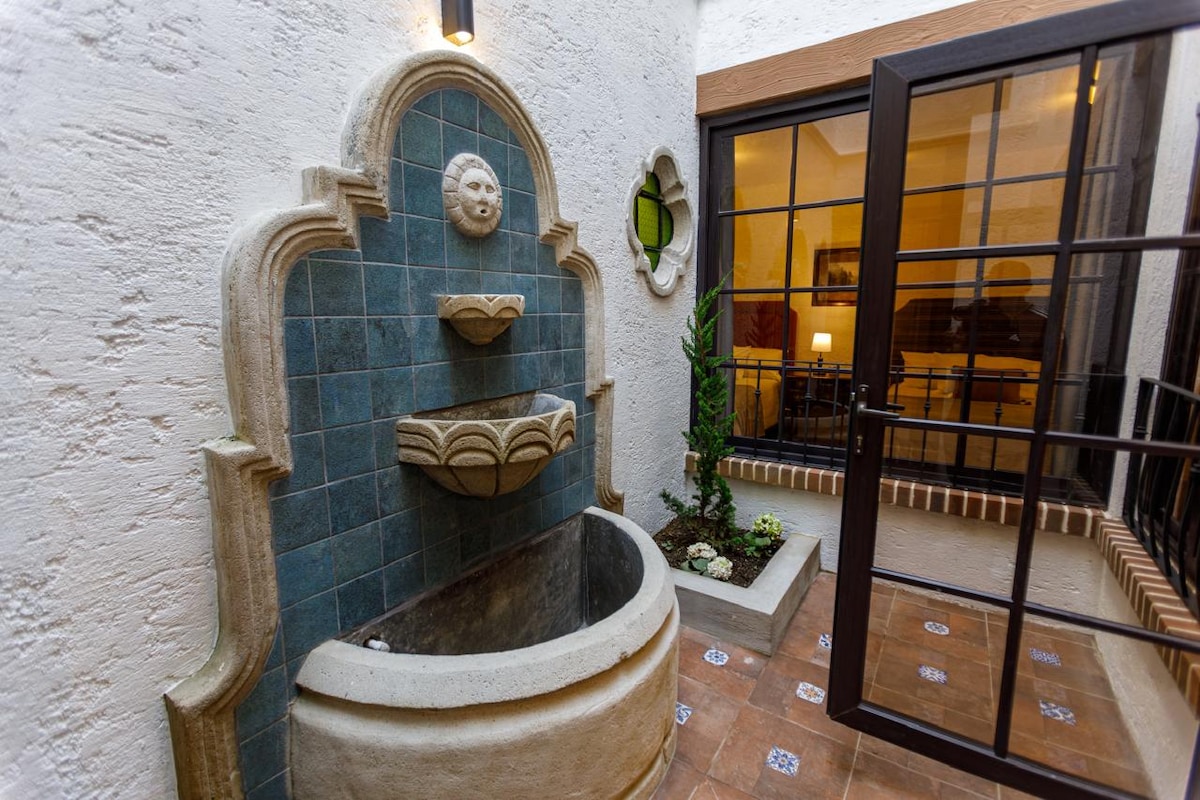 A serene courtyard features a decorative stone fountain with two tiers, complemented by a vibrant blue tiled wall. Surrounding plants add a touch of greenery, while large glass doors provide a view of the inviting interior space. Natural light softly illuminates the area.