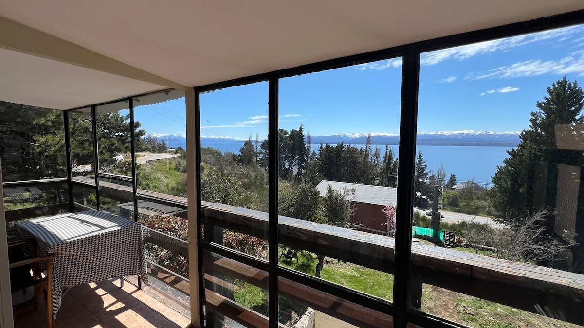 A sunlit balcony features large glass windows offering a panoramic view of the lake and surrounding landscape. A dining table with a checkered tablecloth is positioned near the glass, inviting outdoor meals amidst the scenic backdrop. Lush greenery and distant mountains can be seen beyond.