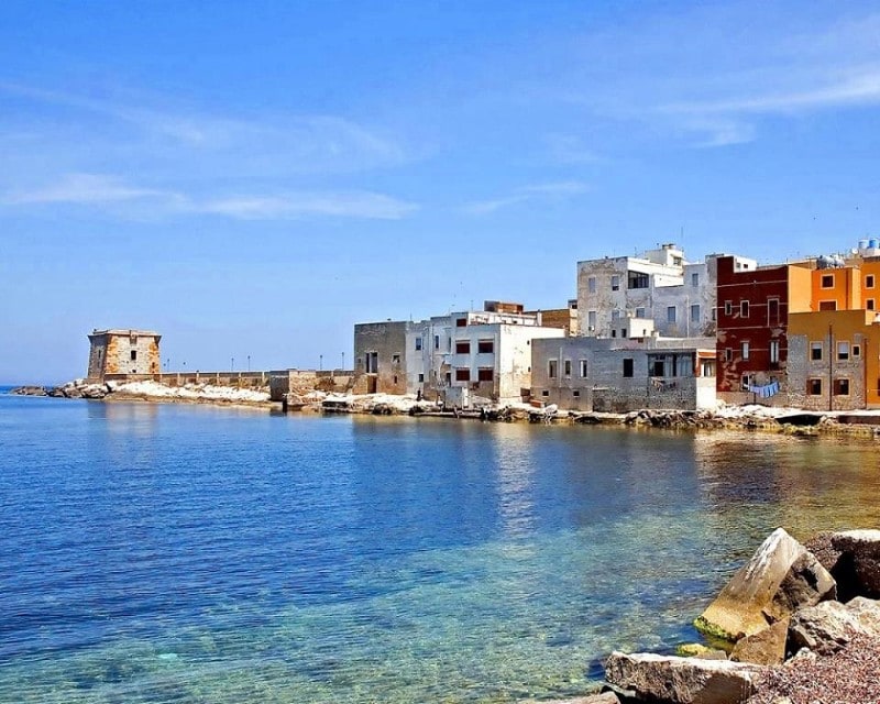 A coastal view reveals a series of buildings lining the waterfront, constructed with light-colored materials. The clear turquoise water reflects the sky, and rocky outcrops are visible in the foreground. A distant tower stands at the edge of the shoreline, adding historical context to the landscape.