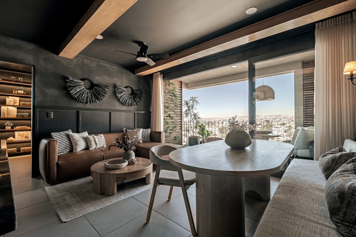 A modern living area features a leather sofa and minimalist decor. A light wooden table is surrounded by four chairs. Large windows offer a panoramic view of the city, while decorative elements add texture to the space. Soft lighting enhances the welcoming ambiance.