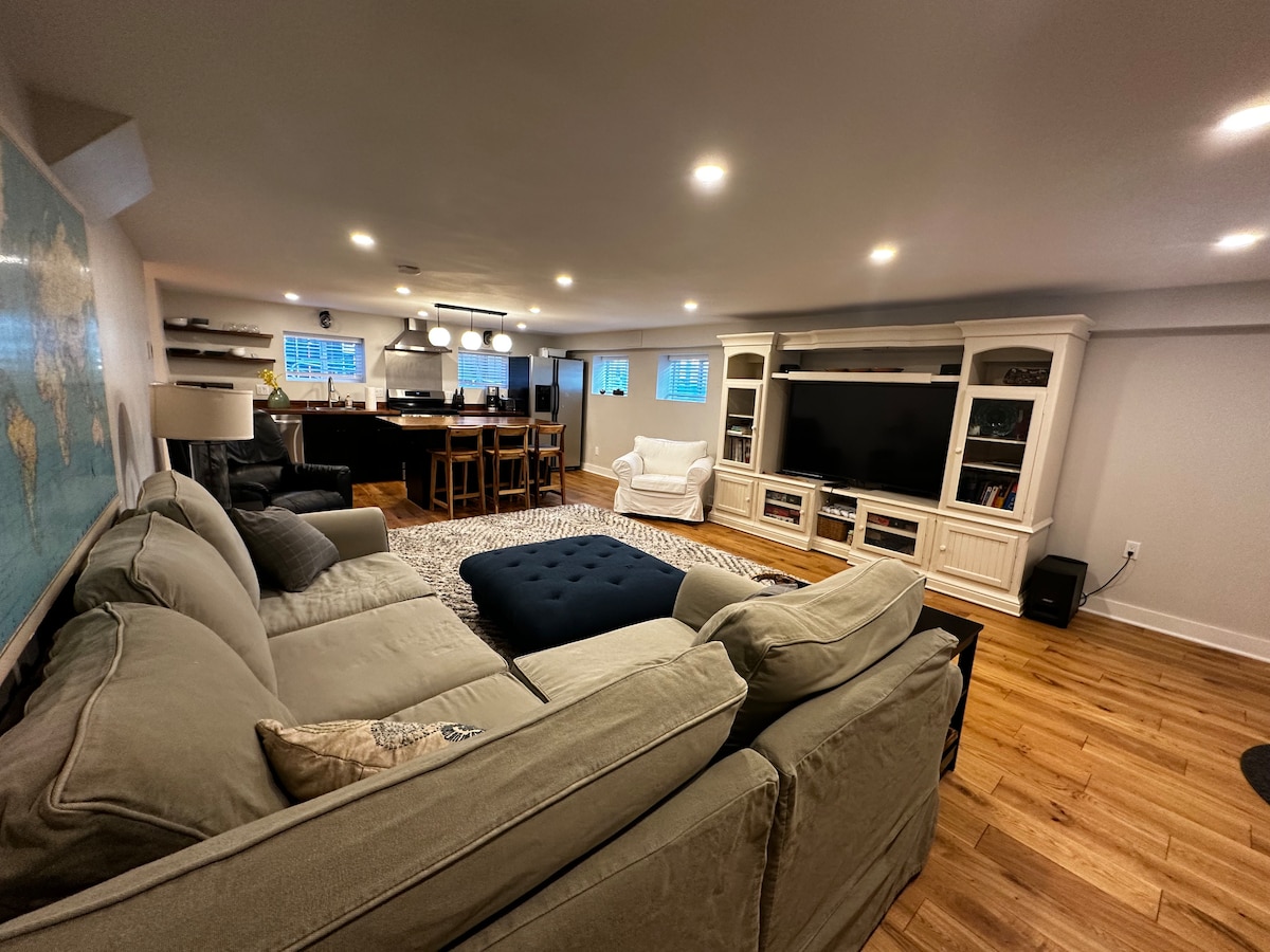 A spacious great room is highlighted by a comfortable sectional sofa, accentuated by a large ottoman. The living area opens into a modern kitchen with bar seating, illuminated by pendant lights. A big screen TV and shelves add functionality to the space, while natural light filters through the windows.