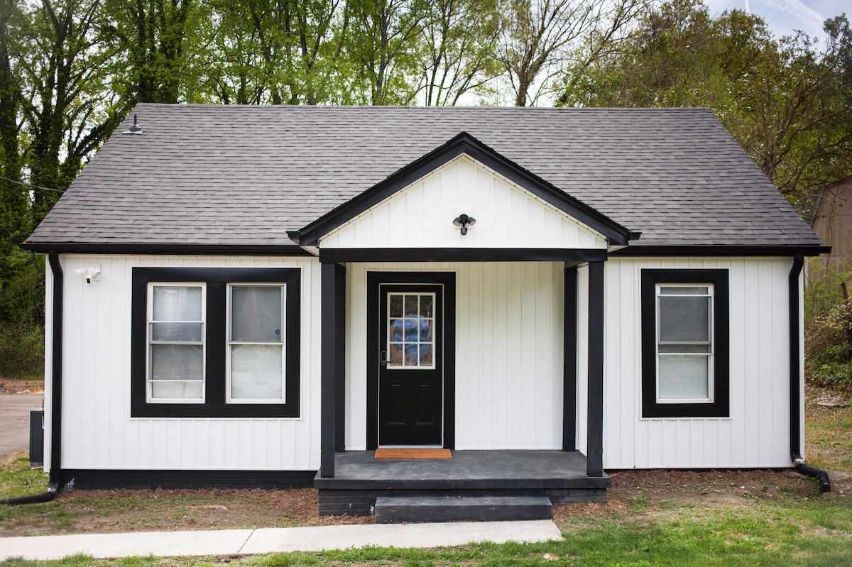 A charming exterior view of a restored villa features a symmetrical design with a central entrance and three front-facing windows. The white siding is contrasted by bold black trim, complemented by a simple porch that leads to a black door, providing an inviting touch.