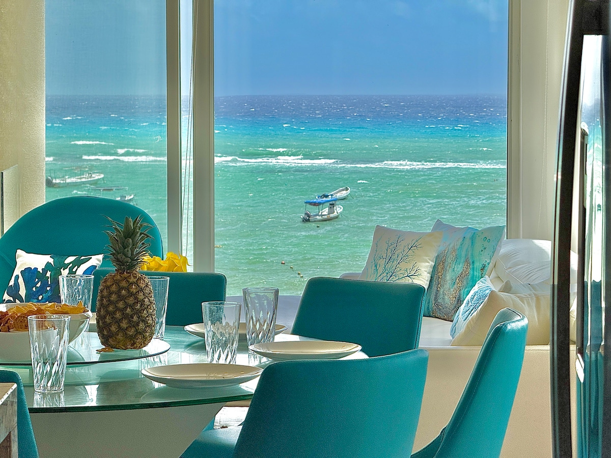 A dining area is visible featuring a glass table surrounded by stylish blue chairs. The ocean can be seen through large windows, with boats gently bobbing on the turquoise water. Decorative pillows are arranged on a nearby couch, adding to the coastal ambiance.