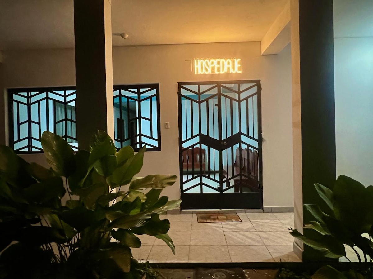 The entrance features a stylish door framed by geometric patterns, with a warm 'HOSPEDAJE' sign illuminated above. Surrounding greenery provides a natural touch, while the tiled floor reflects soft lighting from the adjacent windows.