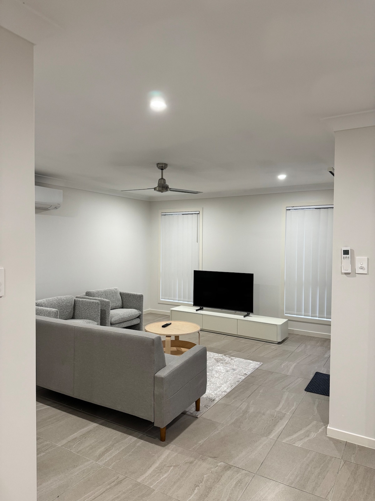 A modern living area is depicted, featuring a grey sectional sofa paired with a small round table. A flat-screen television is positioned on a low white entertainment unit, with two vertical blinds allowing for natural light to enter the space.