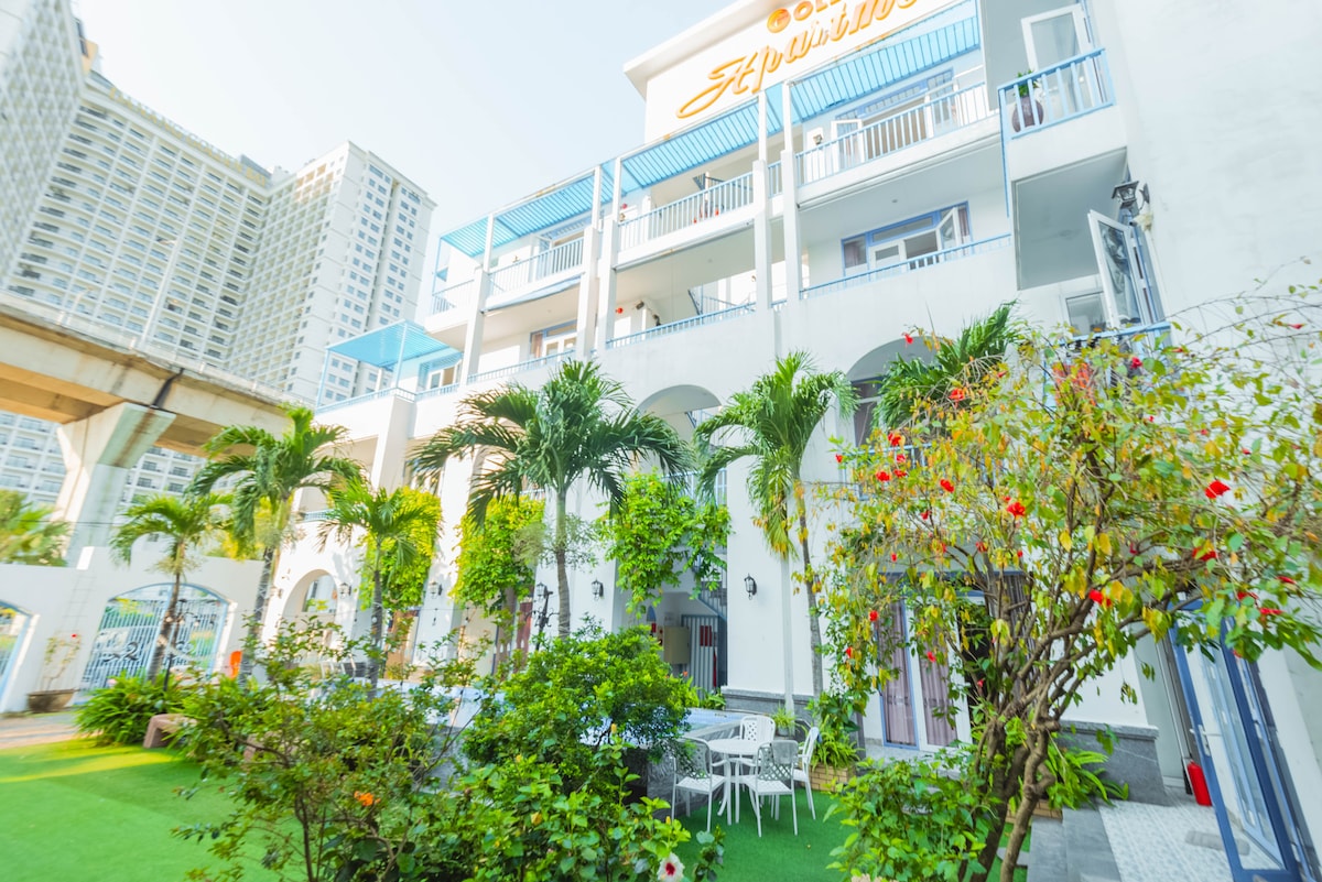 The exterior of the building showcases a three-story structure with a bright blue sign. Lush greenery and colorful flowers surround a grassy area, complemented by white outdoor furniture. The architecture features arches and balconies, contributing to an open and inviting atmosphere.