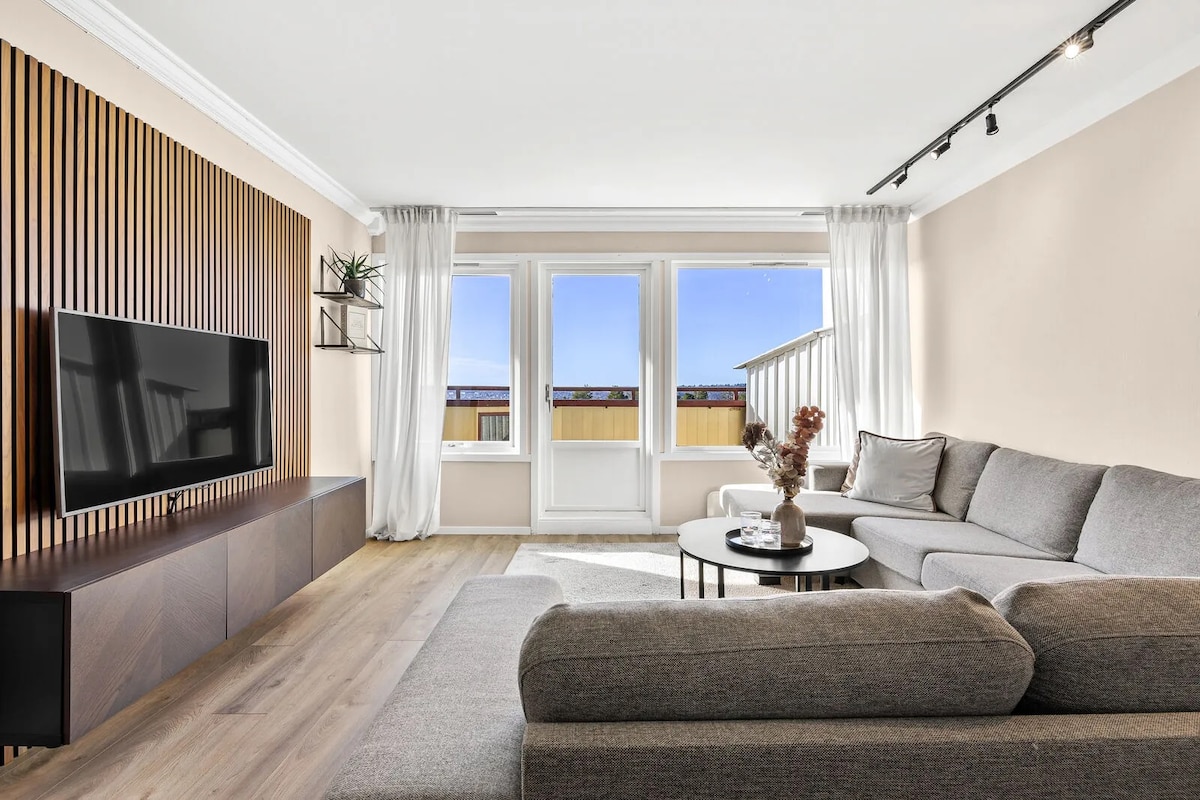 A spacious living area is highlighted by a large sectional sofa and a low coffee table. Natural light pours in through large windows, providing a view of the outside. A wall-mounted television is visible, alongside decorative plants and wooden accents.
