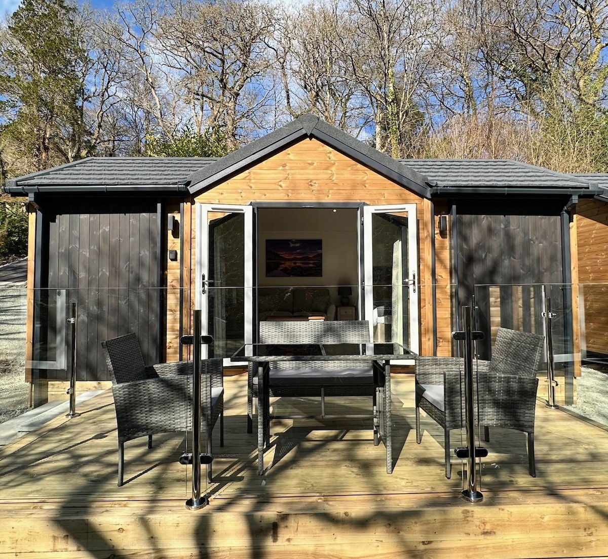 An outdoor decking area includes a table surrounded by four woven chairs, all situated in front of a stylish wooden cabin. The open doors reveal the interior space, while natural surroundings of trees provide a serene backdrop.