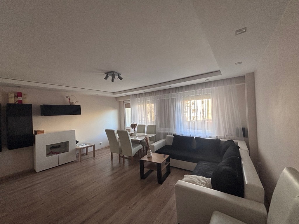 A spacious living area features a combination of light and dark seating arrangements. Floor-to-ceiling windows provide natural light, while a dining table is positioned near a decorative lighting fixture. A mounted television is visible on the wall, complementing the neutral color palette.