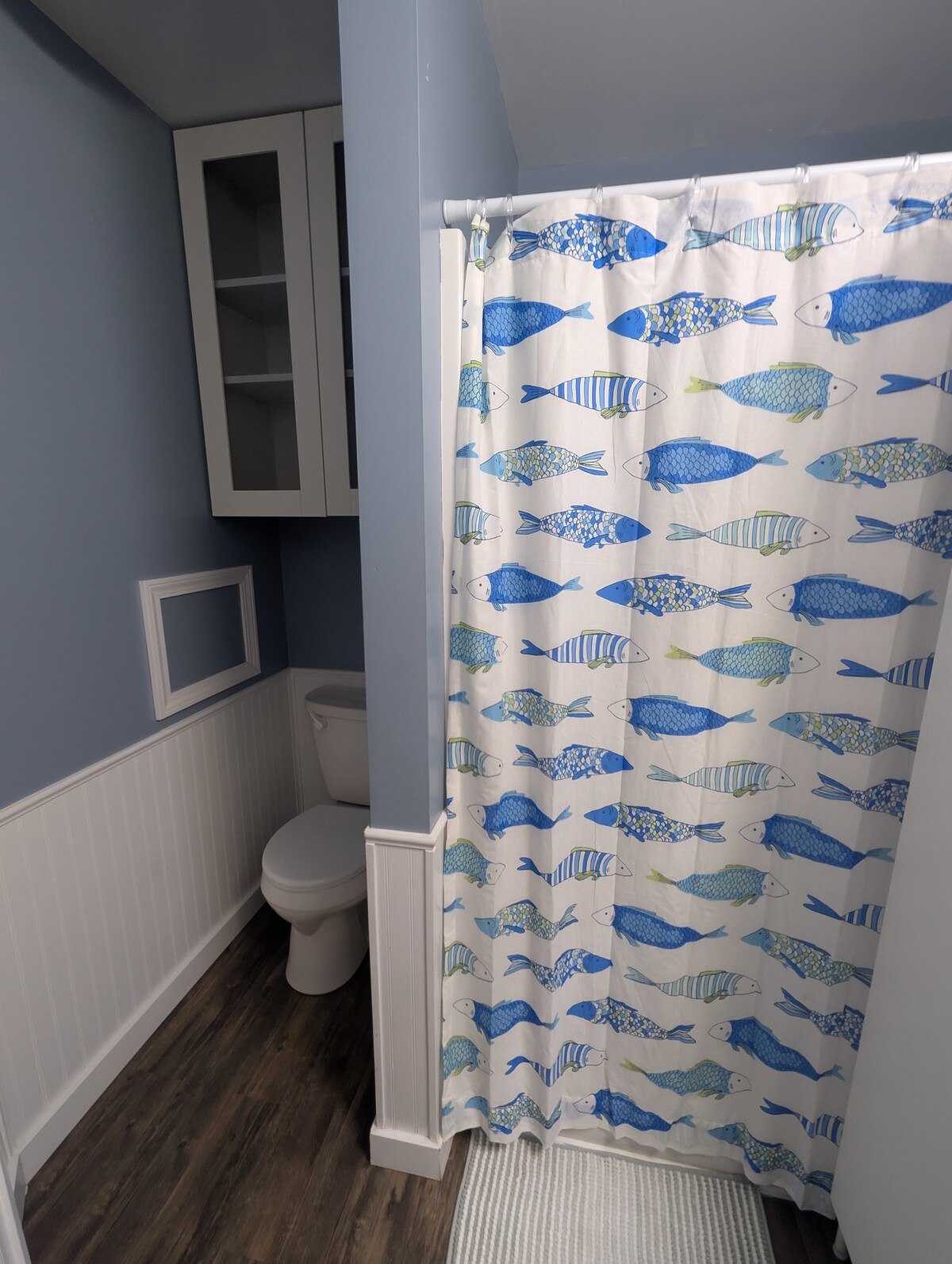 A bathroom is shown with light blue walls and decorative fish-patterned shower curtain. White wainscoting adds texture, while a compact toilet and sink are present. Shelving above the toilet provides extra storage, enhancing functionality.