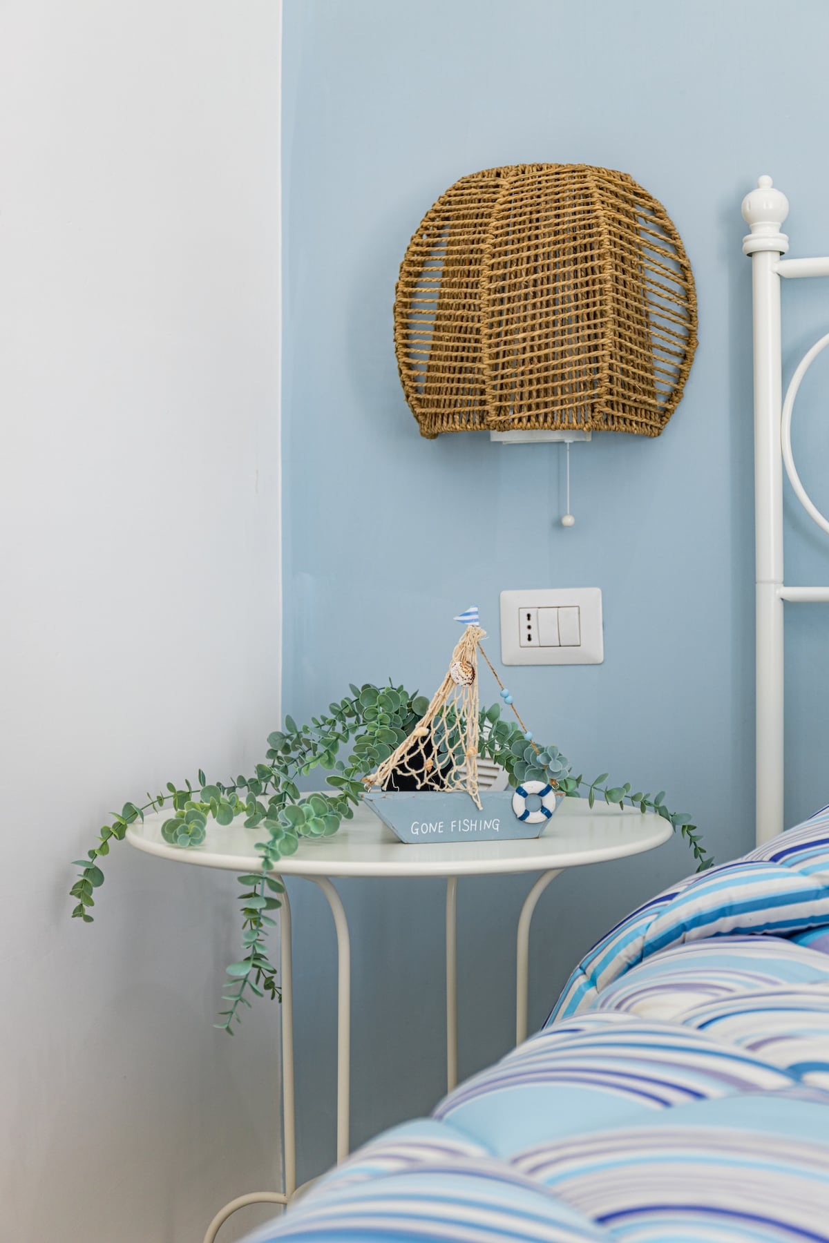 A bedside table is adorned with a decorative fishing-themed figurine and a trailing green plant. A woven wall lamp provides soft lighting, complementing the blue wall and the striped bedding on the nearby bed, creating a calm atmosphere.