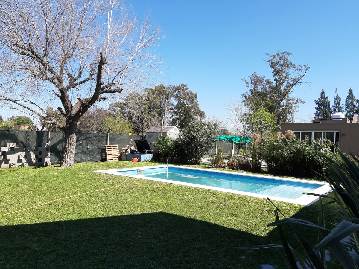 A spacious outdoor area features a well-maintained swimming pool surrounded by lush green grass and trees. A large tree provides shade, and a small patio area is visible in the background, complementing the natural environment.