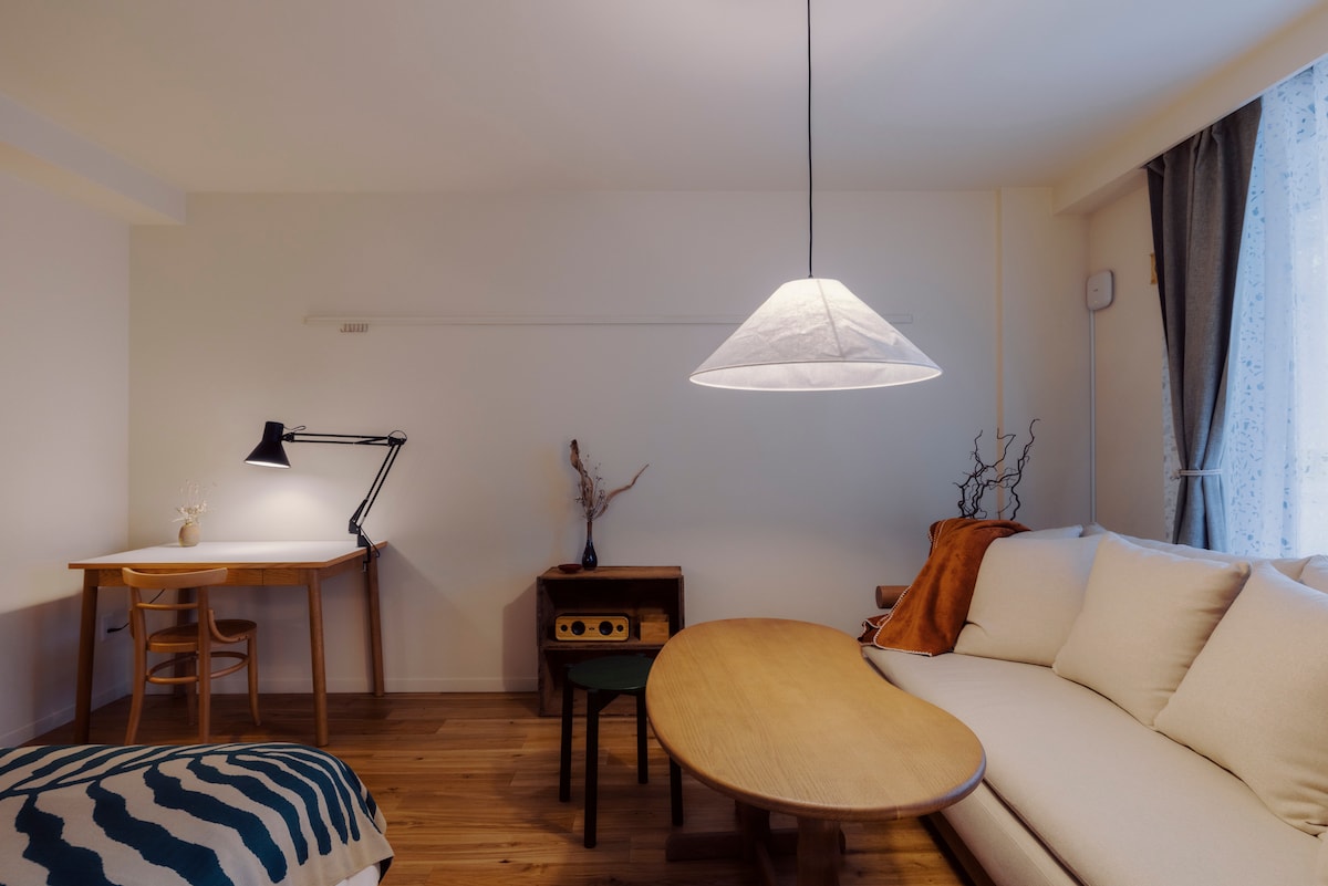 A living area features a light-colored sofa accompanied by a round wooden table. A task lamp and a desk are positioned nearby, while a pendant lamp provides soft illumination overhead. Decorative elements include a small vase and dried branches, enhancing the space's ambiance.