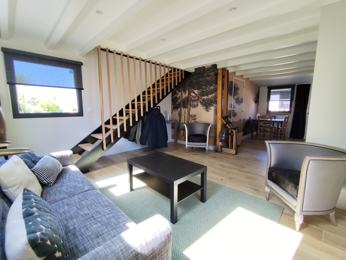A bright living space features a comfortable sofa and stylish armchairs set around a dark coffee table on a light rug. Natural light floods the area, highlighting the wooden staircase and wall art depicting trees. The dining area is visible in the background.