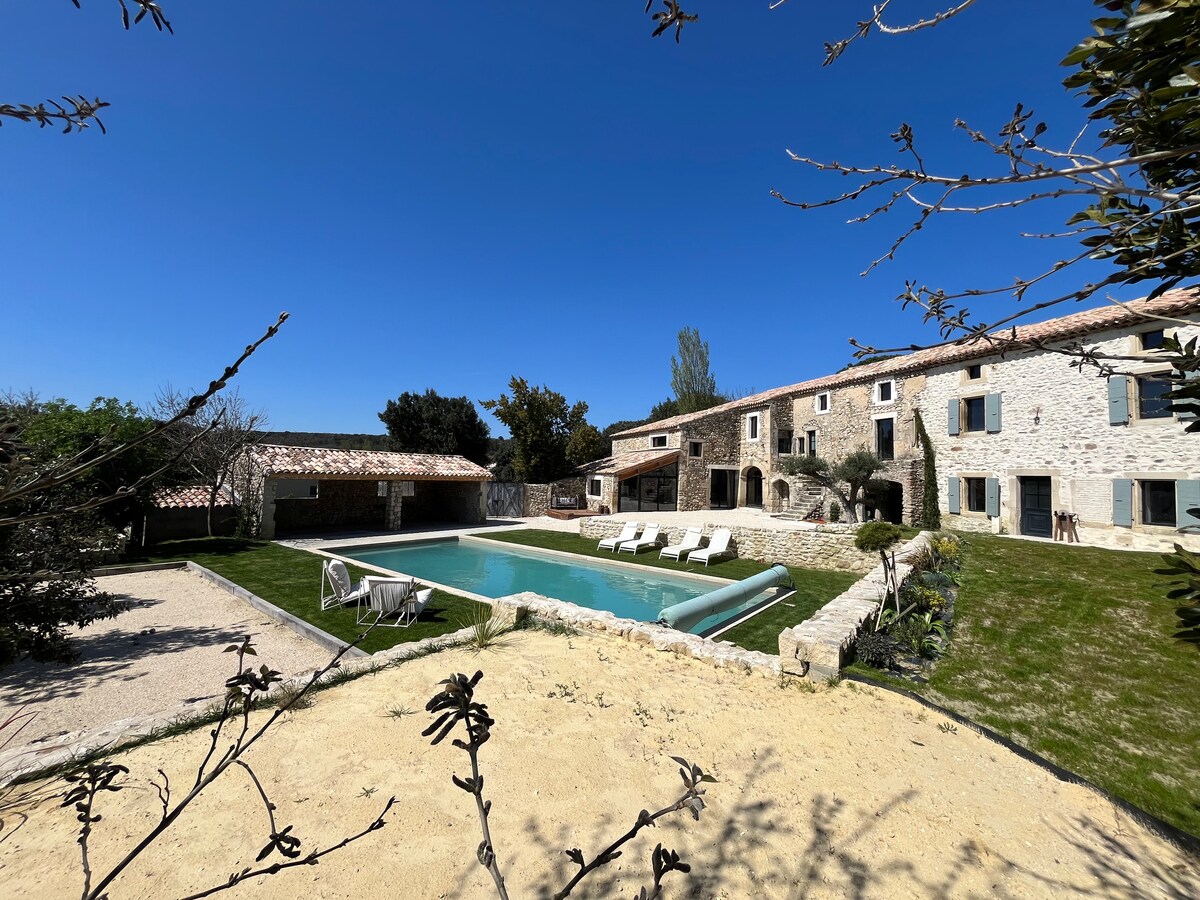 A spacious outdoor area features a private swimming pool surrounded by sun loungers. Stone walls of the renovated house are visible, while well-maintained gardens and trees offer a natural backdrop. A covered area suggests additional outdoor seating and leisure space is available.