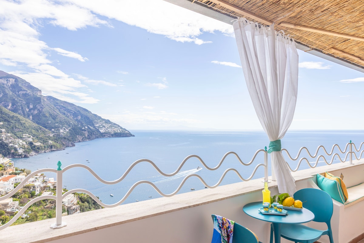 A private terrace offers breathtaking views of the coastline and sea. A small table holds a pitcher and fresh lemons. Blue chairs provide seating, with light fabric curtains gently swaying in the breeze, enhancing the tranquil outdoor space.