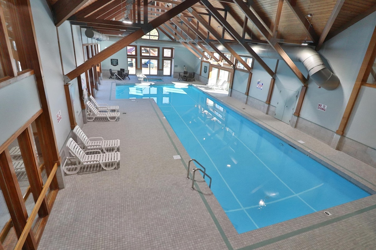An indoor swimming pool is visible beneath a vaulted ceiling with exposed wooden beams. Lounge chairs line the edges of the pool, and large windows allow natural light to brighten the area. A ladder is positioned at one end of the pool for easy access.
